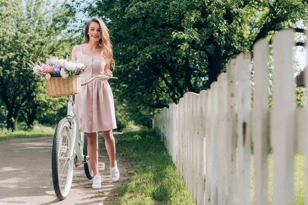 Giovane bella donna in abito con bicicletta retrò con cesto di vimini pieno di fiori in campagna — Foto stock