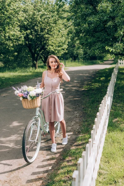Giovane bella donna in abito con bicicletta retrò con cesto di vimini pieno di fiori in campagna — Foto stock