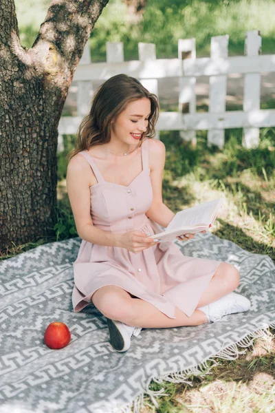Jeune femme souriante lisant le livre tout en se reposant sur la couverture sous l'arbre à la campagne — Photo de stock