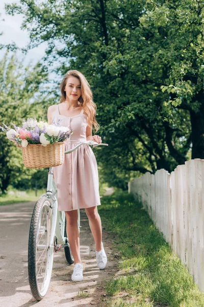 Jeune belle femme en robe avec vélo rétro avec panier en osier plein de fleurs à la campagne — Photo de stock