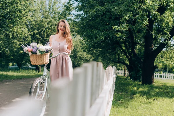 Giovane bella donna in abito con bicicletta retrò con cesto di vimini pieno di fiori in campagna — Foto stock