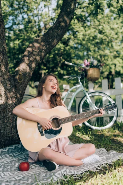 Attraente giovane donna che suona la chitarra acustica mentre riposa sulla coperta sotto l'albero in campagna — Foto stock