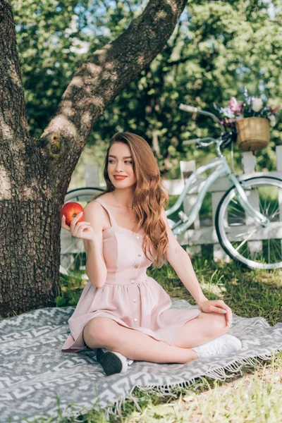 Mujer atractiva sonriente con manzana madura en la mano descansando sobre una manta bajo un árbol en el campo - foto de stock