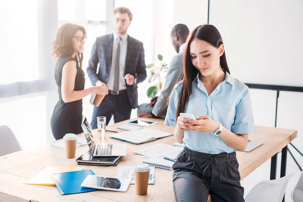 Focalizzazione selettiva della donna d'affari asiatica utilizzando smartphone e colleghi dietro in ufficio — Foto stock