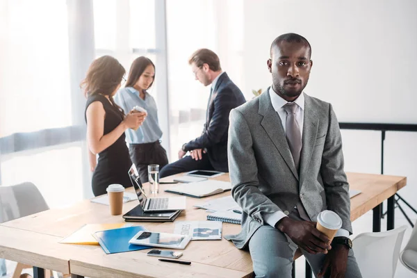 Messa a fuoco selettiva dell'uomo d'affari afroamericano con caffè da andare e colleghi sul posto di lavoro in ufficio — Foto stock