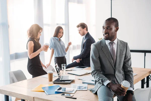 Enfoque selectivo de sonreír hombre de negocios afroamericano con café para llevar y colegas en el lugar de trabajo en la oficina - foto de stock