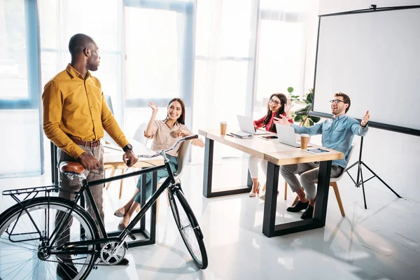 Multicultural business people greeting african american colleague with bicycle in office — Stock Photo