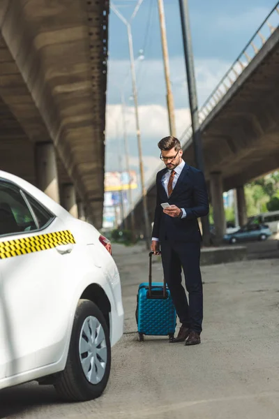 Joven guapo usando el teléfono inteligente mientras está de pie con la maleta cerca de taxi - foto de stock