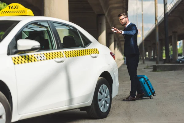 Стильний молодий чоловік зі смартфоном і валіза, дивлячись на таксі — стокове фото