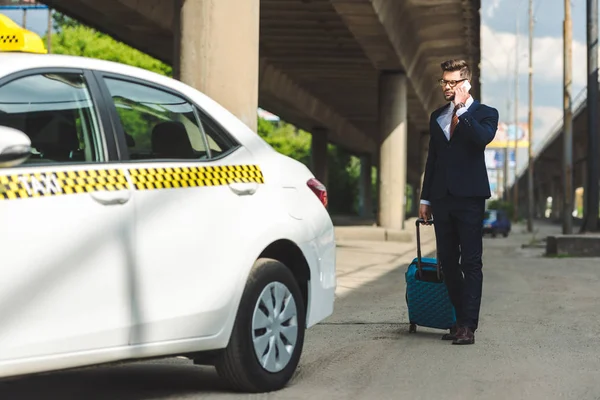 Handsome young man in suit and eyeglasses talking by smartphone while standing with suitcase near taxi — Stock Photo