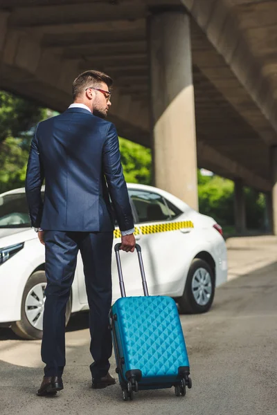 Vista trasera de joven hombre de negocios con estilo con la maleta de pie cerca de taxi - foto de stock