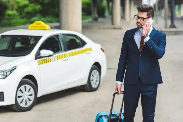 Hombre guapo hablando por teléfono inteligente mientras está de pie con la maleta en taxi - foto de stock