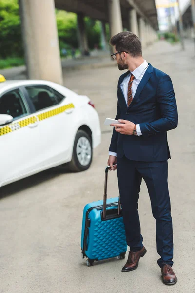 Bell'uomo con valigia e smartphone guardando taxi in strada — Foto stock