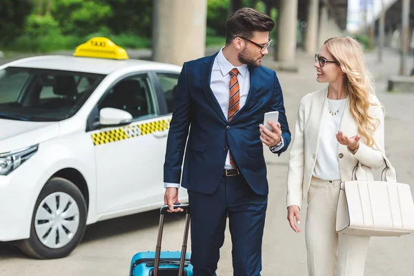 Bella felice giovane coppia in abito formale sorridendo l'un l'altro mentre si va con valigia vicino taxi — Foto stock