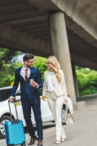 Feliz jovem casal sorrindo uns aos outros enquanto vai com mala perto de táxi — Fotografia de Stock