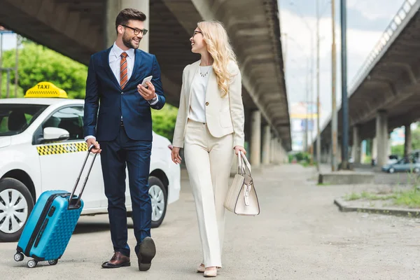 Stylish young couple smiling each other while going with suitcase near taxi — Stock Photo