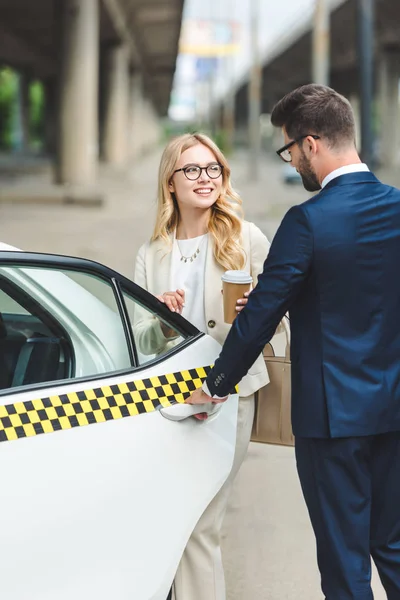 Belle fille blonde souriante tenant du café pour aller et regardant bel homme ouvrir la porte du taxi — Photo de stock