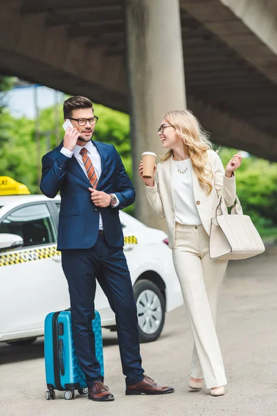 Chica rubia sonriente sosteniendo taza de papel y mirando al hombre guapo hablando por teléfono inteligente mientras está de pie con la maleta cerca de taxi - foto de stock