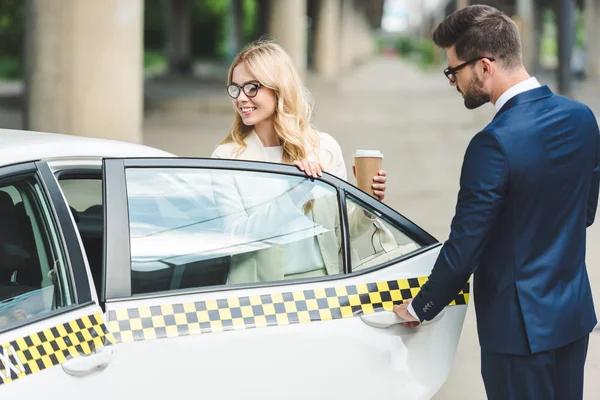 Bell'uomo che apre la portiera dell'auto mentre sorride donna bionda con tazza di carta seduta in taxi — Foto stock