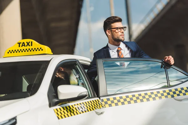 Hansome joven con traje y anteojos mirando hacia otro lado mientras está sentado en taxi - foto de stock