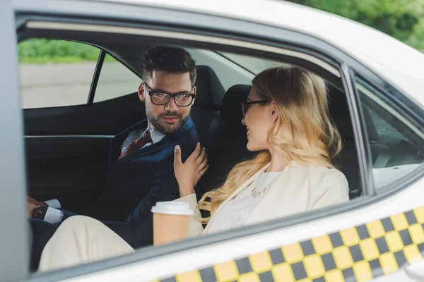 Smiling young woman holding paper cup and flirting with handsome man sitting in taxi — Stock Photo