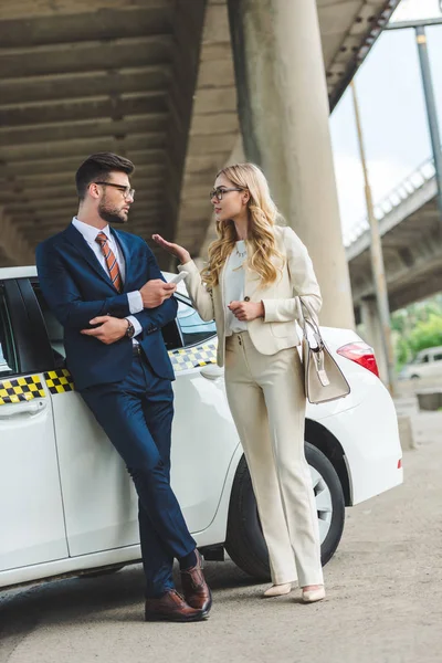 Young couple in formal wear and eyeglasses talking and looking at each other while standing near taxi — Stock Photo