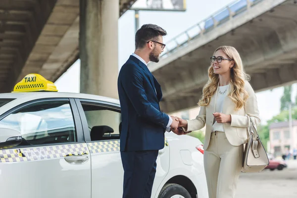 Stylish young couple in eyeglasses shaking hands and smiling each other while standing together near taxi cab — Stock Photo