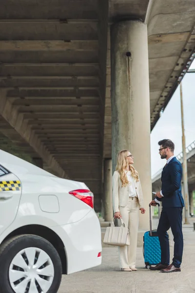 Elegante pareja joven hablando y mirándose mientras está de pie con la maleta cerca de taxi - foto de stock