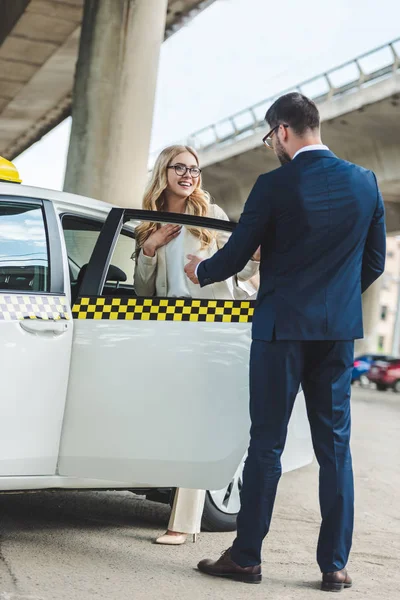 Hombre de traje abriendo la puerta del coche a una hermosa mujer sonriente sentada en taxi - foto de stock