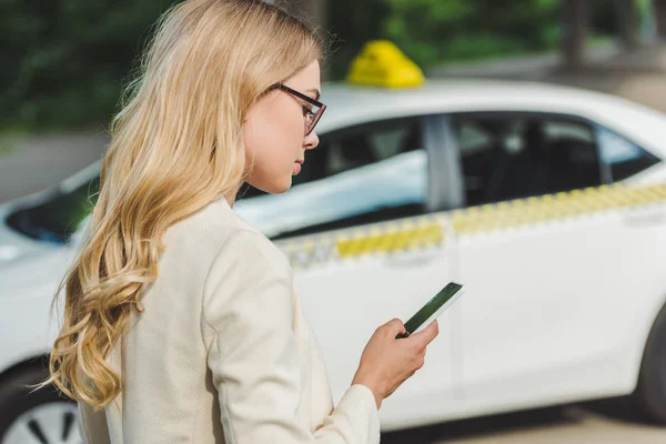Beautiful blonde woman in eyeglasses using smartphone while standing near taxi — Stock Photo