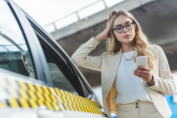 Hermosa chica rubia de moda en gafas inclinadas en taxi cabina y el uso de teléfono inteligente - foto de stock