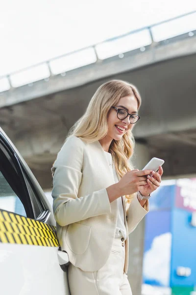 Beautiful smiling blonde woman in eyeglasses leaning at taxi cab and using smartphone — Stock Photo