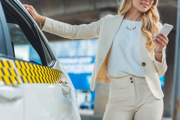 Plan recadré de fille blonde souriante à l'aide d'un smartphone tout en se tenant près de taxi — Photo de stock