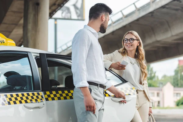 Jeune homme ouvrant la porte du taxi et regardant souriant femme blonde avec smartphone — Photo de stock