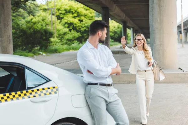 Sorridente giovane donna con gli occhiali agitando mano all'uomo bello appoggiato alla macchina del taxi — Foto stock