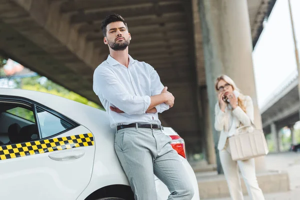 Vista de ángulo bajo del joven con los brazos cruzados apoyados en el coche de taxi y mirando a la cámara mientras la mujer de pie detrás - foto de stock