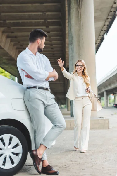 Smiling young woman in eyeglasses waving hand to man leaning at car — Stock Photo