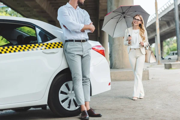 Cropped shot of male driver leaning at cab while smiling woman with umbrella going to the taxi — Stock Photo