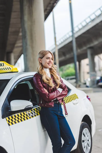 Beautiful blonde girl standing with crossed arms near taxi cab and looking away — Stock Photo