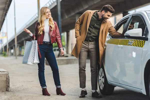 Lächelnde junge Frau hält Einkaufstüten in der Hand und blickt ihren glücklichen Freund an, der ins Taxi blickt — Stockfoto