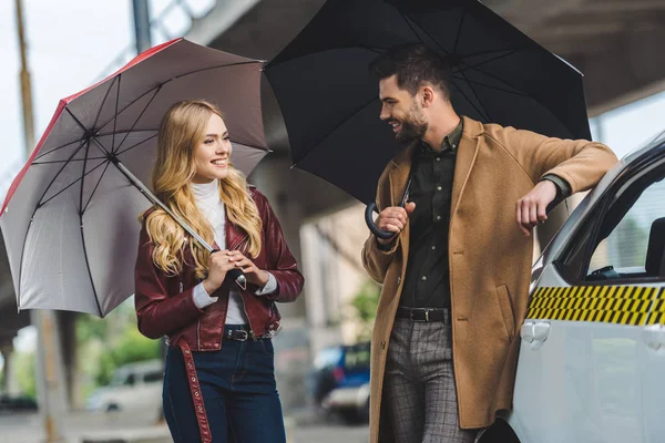 Glückliches junges Paar hält Regenschirme in der Hand und lächelt einander an, während es im sauberen Taxiauto zusammensteht — Stockfoto