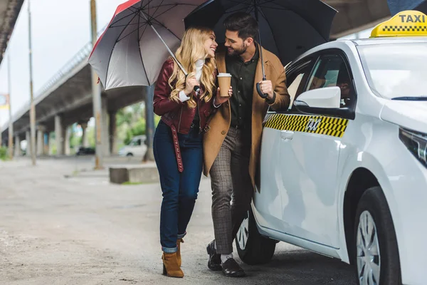 Счастливая молодая пара с зонтиками, улыбающимися друг другу, стоя возле такси — стоковое фото