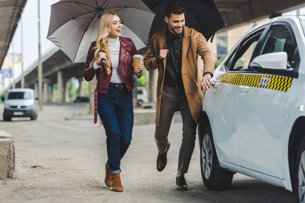 Smiling young couple with umbrellas running to taxi cab — Stock Photo