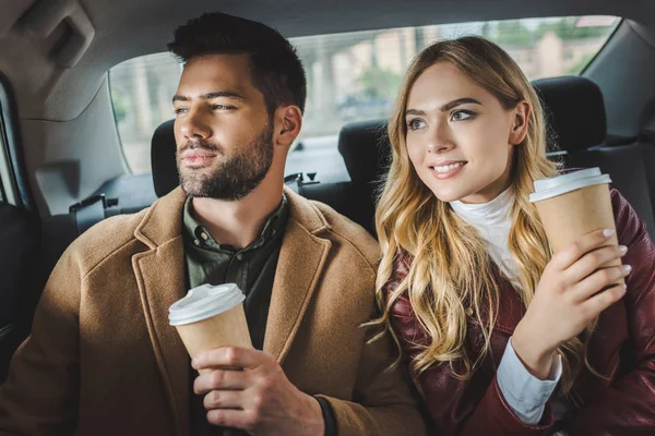 Sonriente elegante pareja joven con vasos de papel sentados juntos en taxi - foto de stock