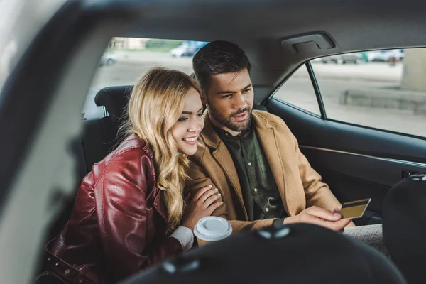 Sonriente pareja joven con taza de papel y tarjeta de crédito sentado en taxi - foto de stock