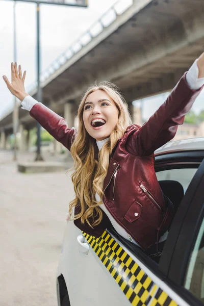 Fröhliches blondes Mädchen hebt die Hände und schaut durch das Taxifenster weg — Stockfoto