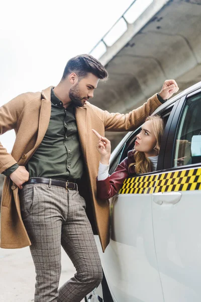 Serious young woman sitting in taxi and pointing with finger at man leaning at car — Stock Photo