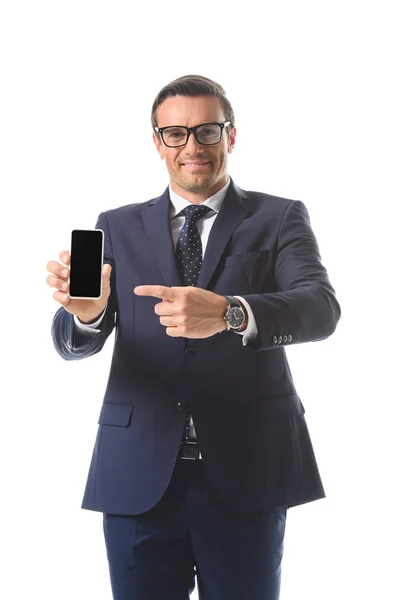 Smiling businessman in eyeglasses pointing on smartphone with blank screen isolated on white background — Stock Photo