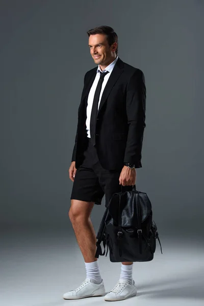 Smiling stylish man in shorts holding backpack on grey background — Stock Photo