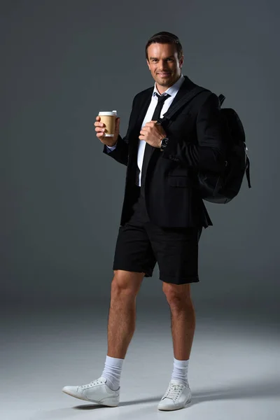Hombre con estilo feliz en pantalones cortos con mochila sosteniendo taza de papel de café sobre fondo gris - foto de stock